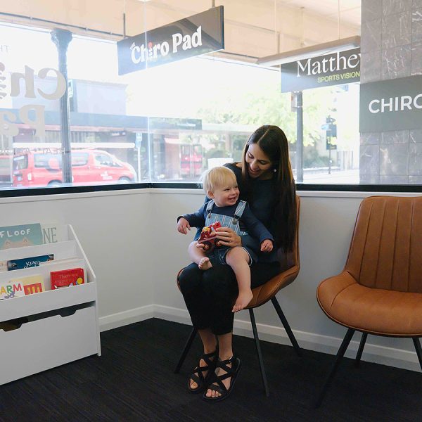 Woman and toddler sitting together in a chiropractic clinic waiting area with children’s books nearby.